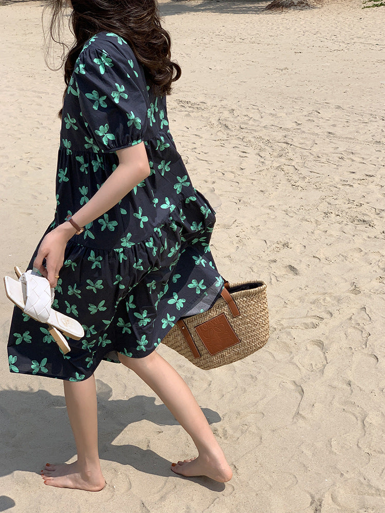 A woman walks along a beach wearing a black dress with a green floral print and carrying a straw bag and white sandals.