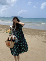 A woman stands on a beach holding a bouquet of flowers and a straw bag, covering her eyes with her hand.