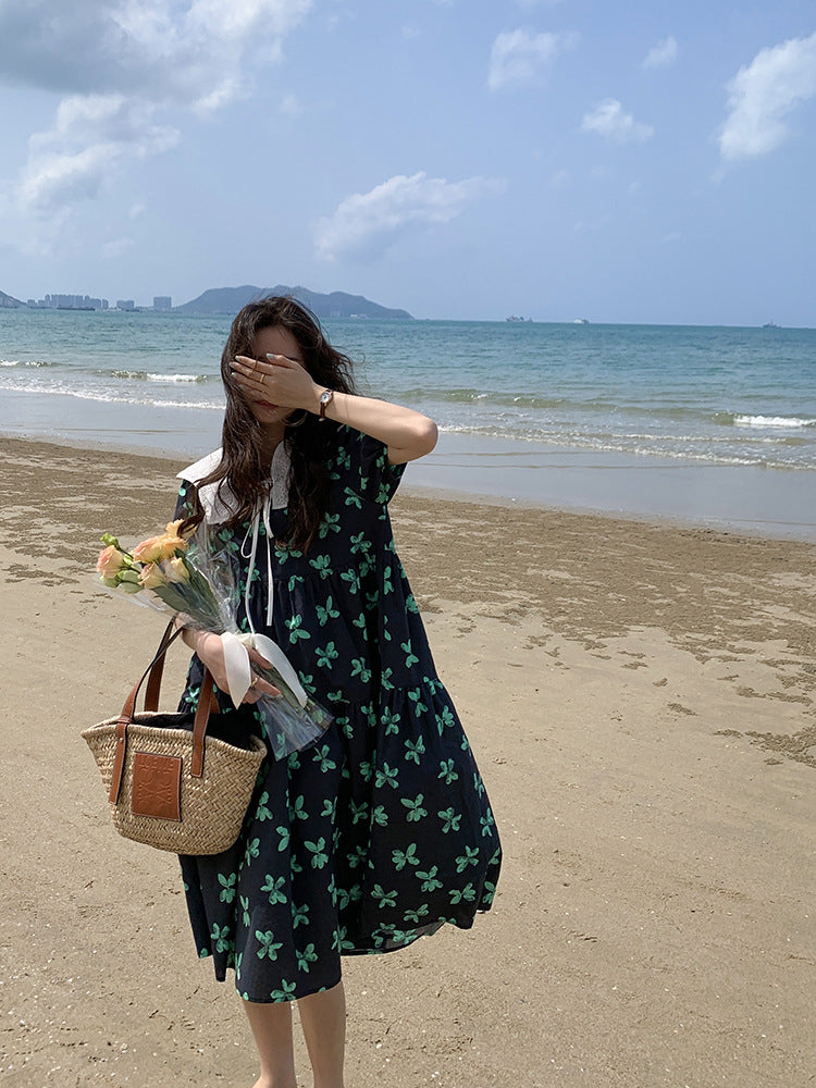 A woman stands on a beach holding a bouquet of flowers and a straw bag, covering her eyes with her hand.