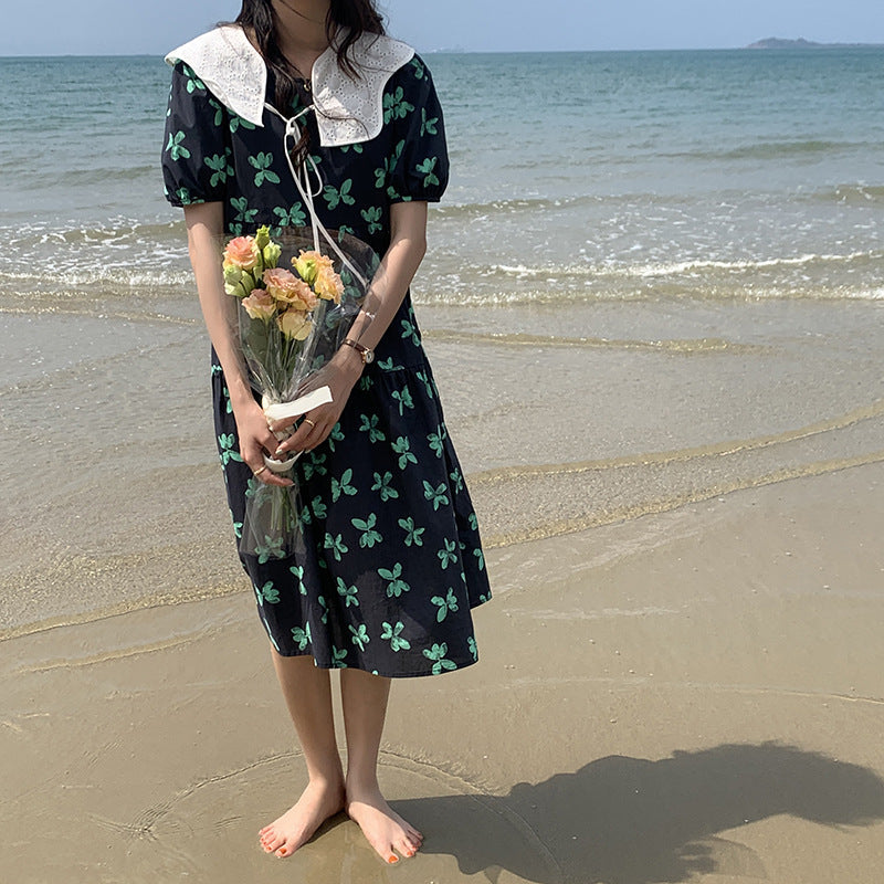 A woman stands on a beach holding a bouquet of flowers while wearing a black dress with a green floral print and a white lace collar.