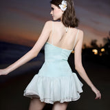 Back view of a woman wearing a light blue and white ruffled dress with spaghetti straps on a beach at sunset.