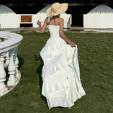A woman in a white strapless ruffled maxi dress with an asymmetrical hem and high slit turns away from the camera. She wears a wide-brimmed straw hat.