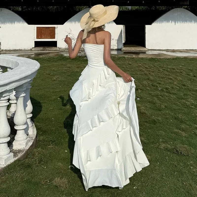 A woman in a white strapless ruffled maxi dress with an asymmetrical hem and high slit turns away from the camera. She wears a wide-brimmed straw hat.