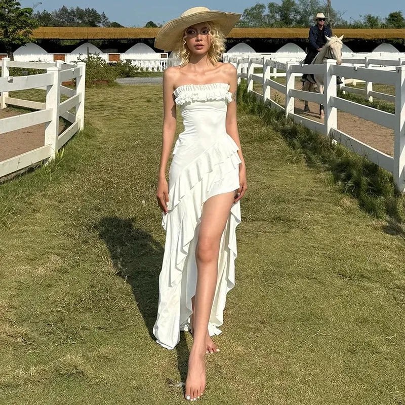 A woman in a white strapless ruffled maxi dress with an asymmetrical hem and high slit walks down a grassy path with a white fence on either side. She wears a wide-brimmed straw hat.
