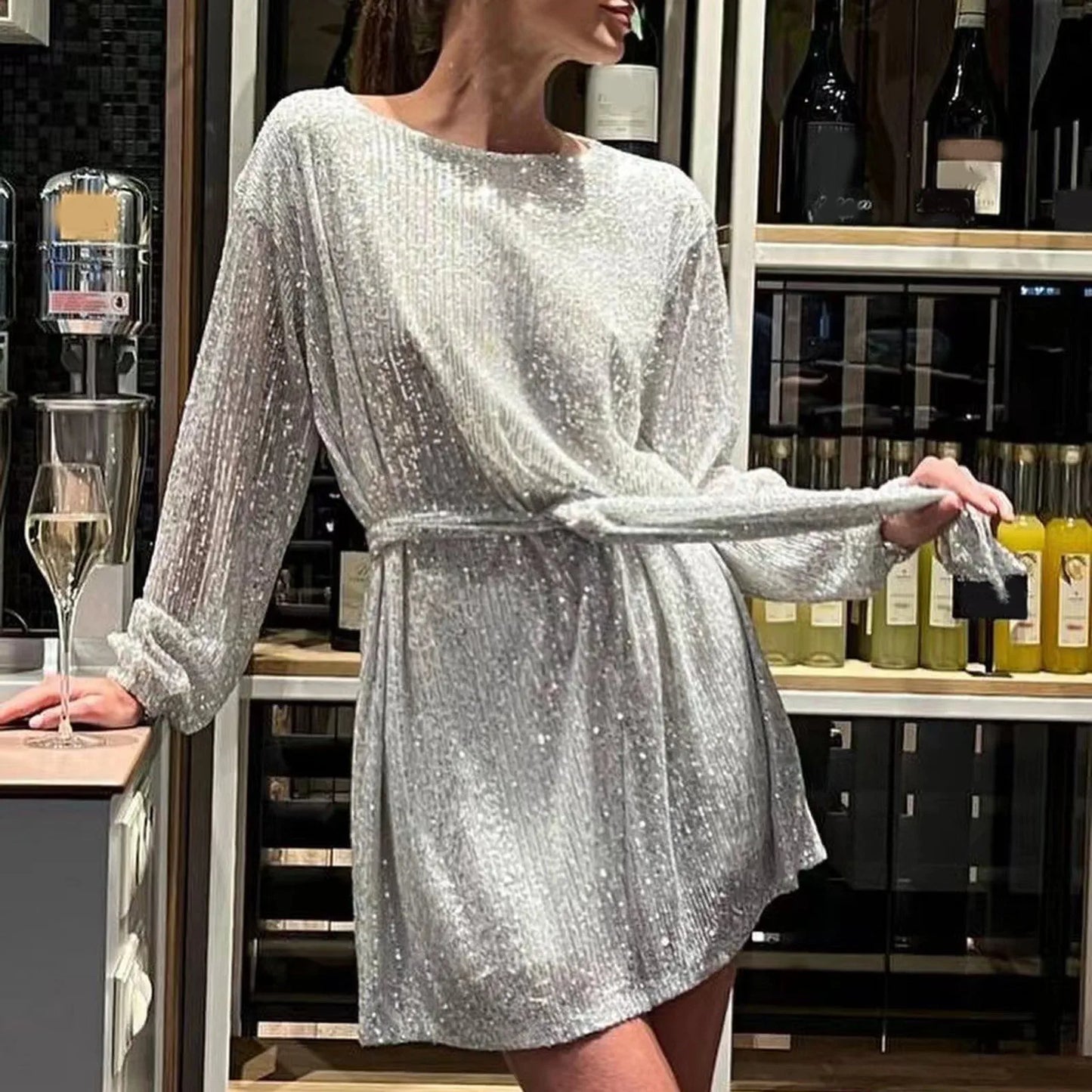 Woman wearing a silver sequined dress with long sleeves and a belted waist, standing in a bar.