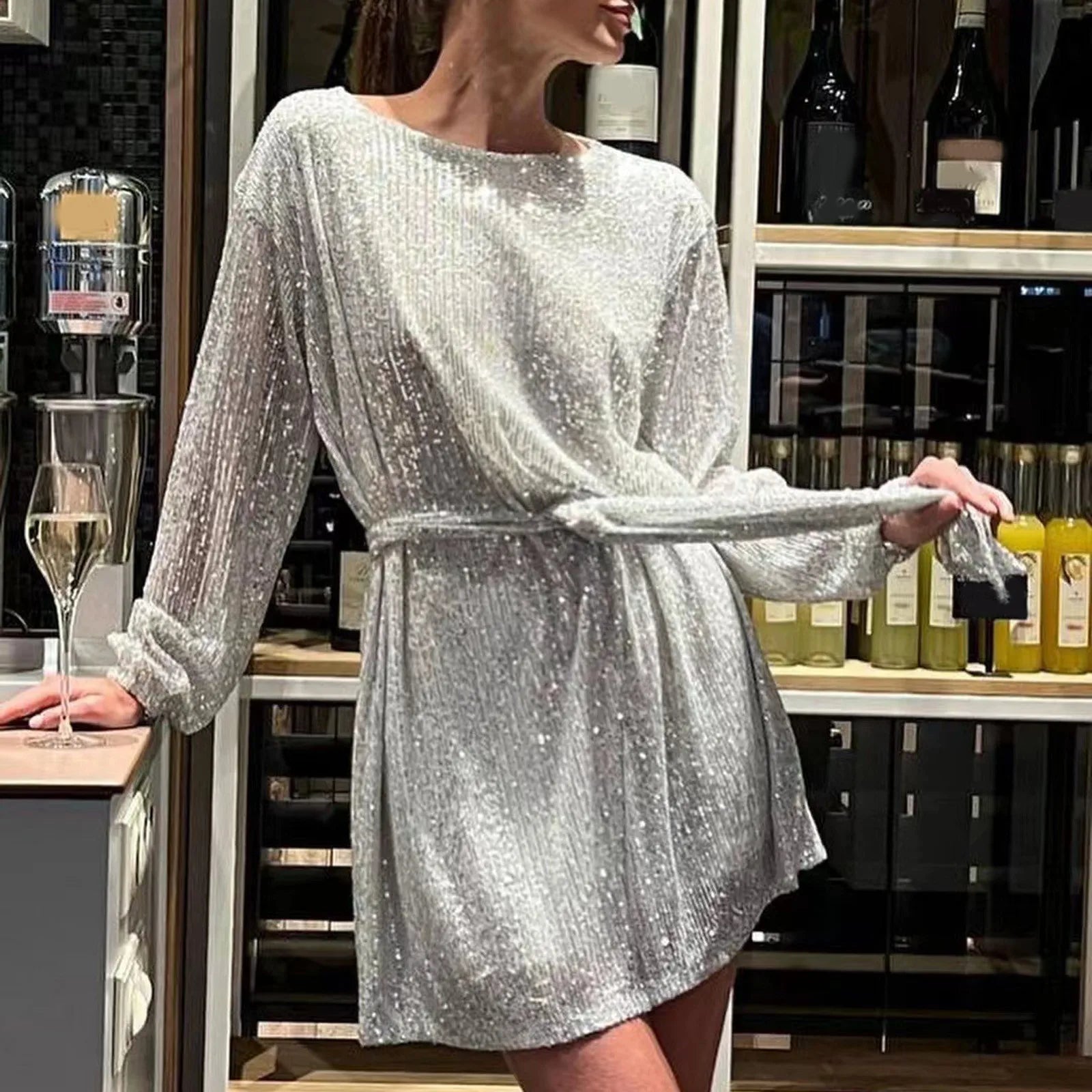 Woman wearing a silver sequined dress with long sleeves and a belted waist, standing in a bar.