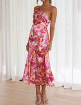A woman in a pink and orange floral print one-shoulder satin dress lifts the hem slightly.