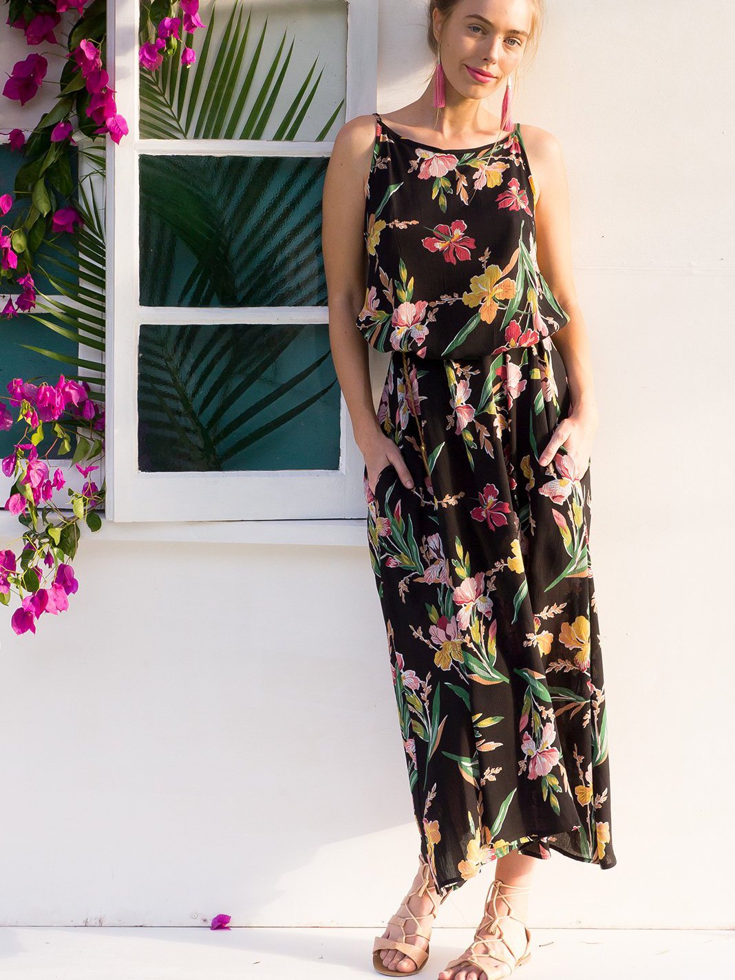 Woman in a black floral maxi dress with her hands in her pockets, leaning against a white wall.