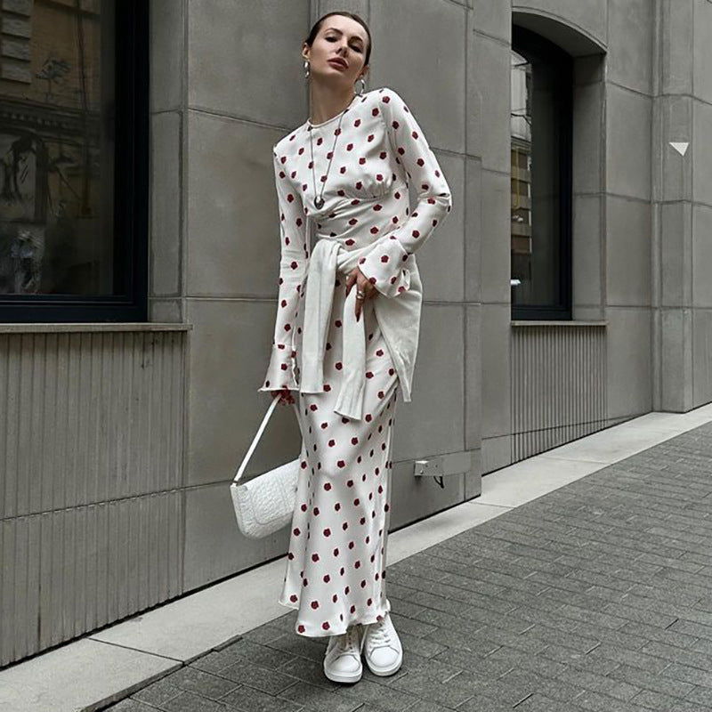 Woman wearing a white polka dot maxi dress with red polka dots and bell sleeves, with white sneakers and a white sweater tied around her waist.
