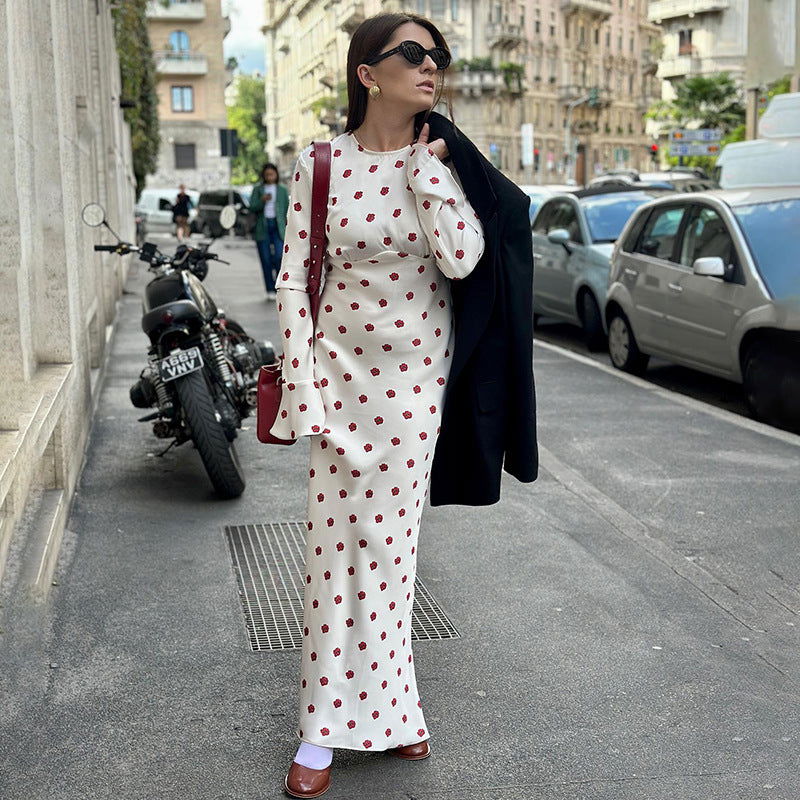 Woman wearing a white polka dot maxi dress with red polka dots and bell sleeves, carrying a black coat over her shoulder and a red bag.