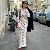 Woman wearing a white polka dot maxi dress with red polka dots and bell sleeves, carrying a black coat over her shoulder and a red bag.
