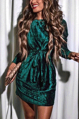 Woman in a long-sleeved emerald green velvet mini dress with a tie waist and mock neck, holding a gold clutch.