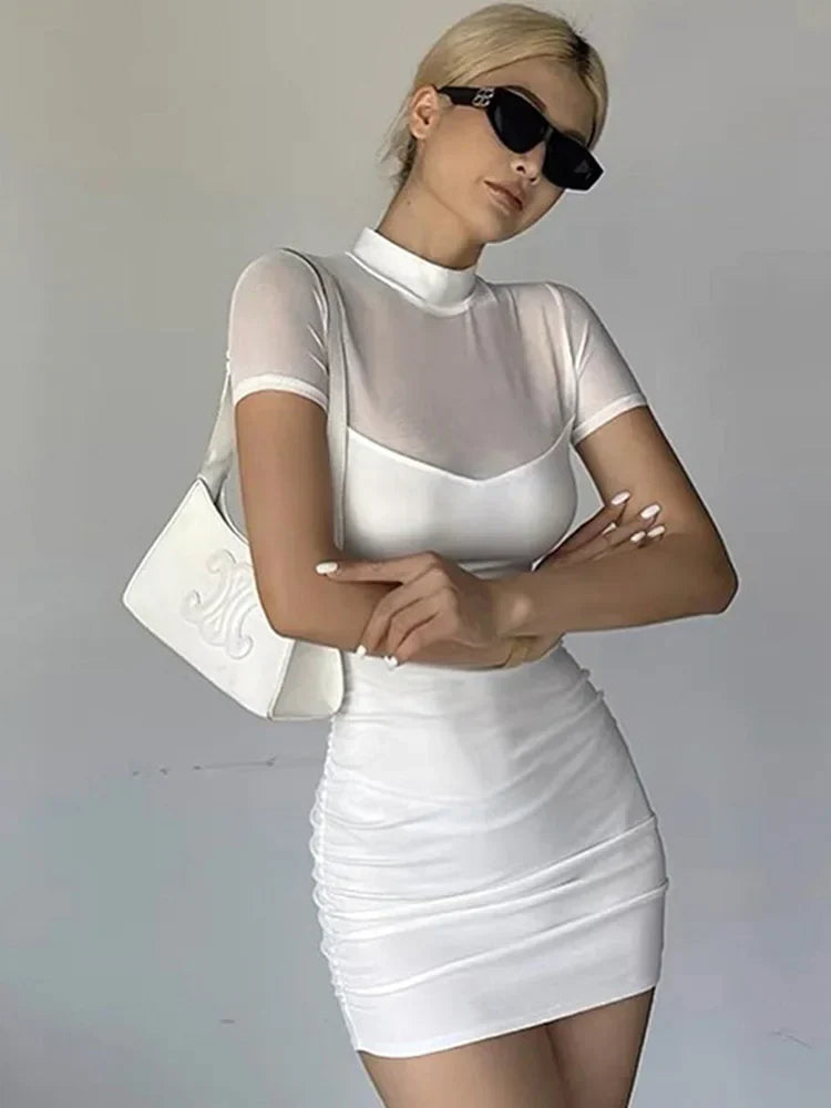 A woman in a white ruched dress with a sheer neckline, mock neck, and short sleeves, holding a white handbag.