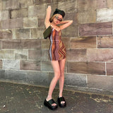 A young woman stands in front of a brick wall wearing a brown and orange abstract print mini dress, black platform sandals, sunglasses, and a black beanie. She has a small brown shoulder bag.
