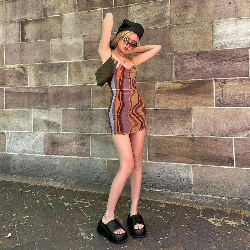 A young woman stands in front of a brick wall wearing a brown and orange abstract print mini dress, black platform sandals, sunglasses, and a black beanie. She has a small brown shoulder bag.