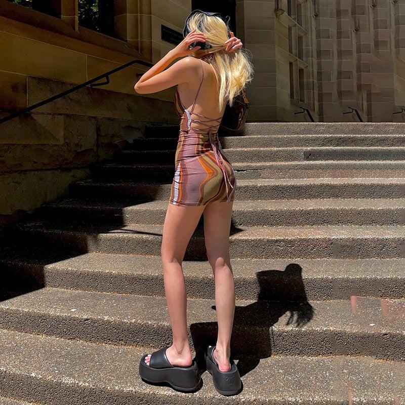 A young woman from behind stands on stone stairs wearing a brown and orange abstract print mini dress, black platform sandals, and sunglasses.