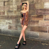 A young woman stands in front of a brick wall wearing a brown and orange abstract print mini dress, black platform sandals, sunglasses, and a black beanie.