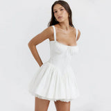 Woman wearing a white mini dress with spaghetti straps, a sweetheart neckline, and a pleated skirt with lace trim, with hands behind back.