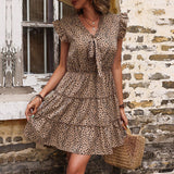 A woman wearing a tan and black leopard print tiered mini dress with ruffle sleeves and a bow tie neckline, holding a straw bag.