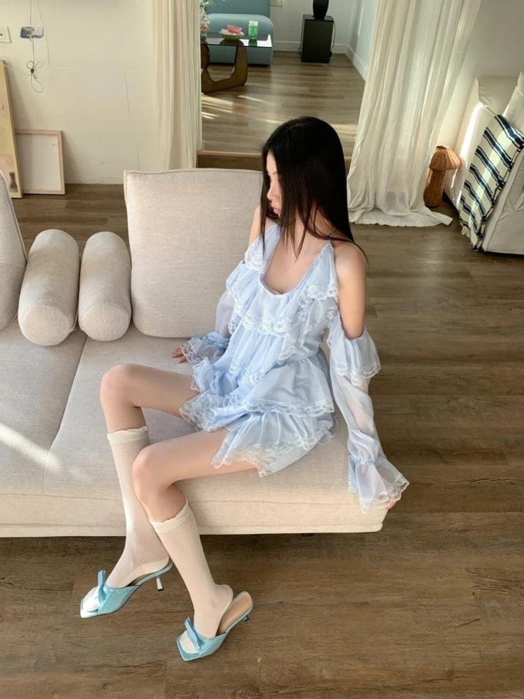 A young woman sits on a couch wearing a light blue, ruffled, off-the-shoulder dress with lace trim, white socks, and blue slip-on shoes.