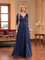 Woman in a navy blue sparkly A-line evening gown with a plunging V-neck and sheer three-quarter sleeves, standing in a room with a couch and a table with lamps.