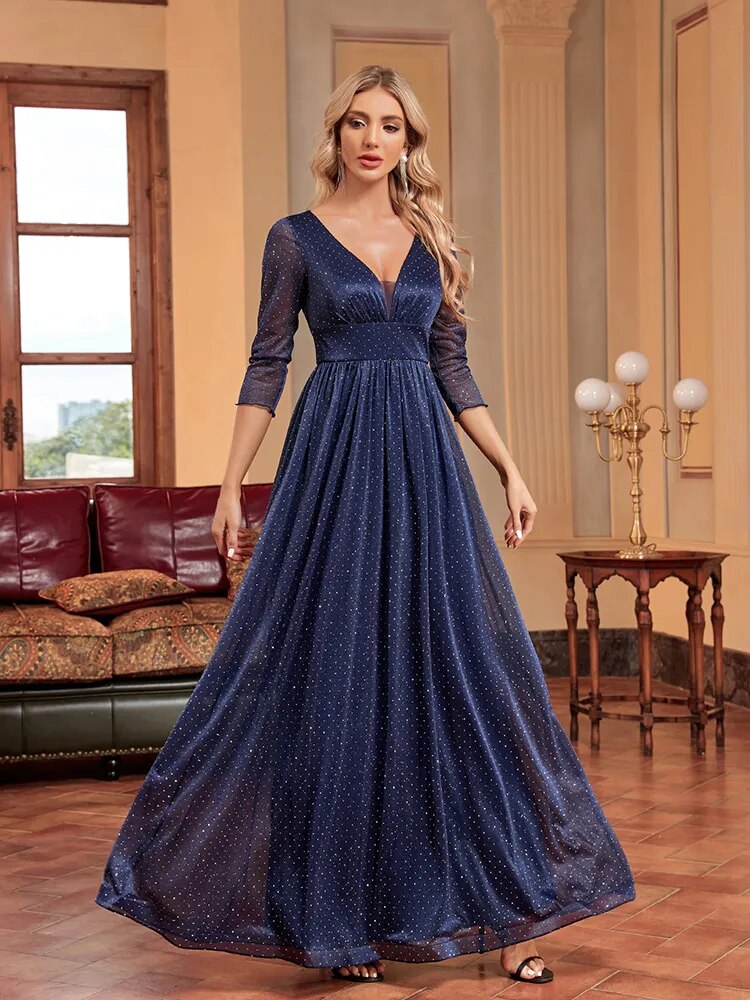 Woman in a navy blue sparkly A-line evening gown with a plunging V-neck and sheer three-quarter sleeves, standing with her hands by her side.