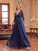 Woman in a navy blue sparkly A-line evening gown with a plunging V-neck and sheer three-quarter sleeves, posing with her hand on her hip.