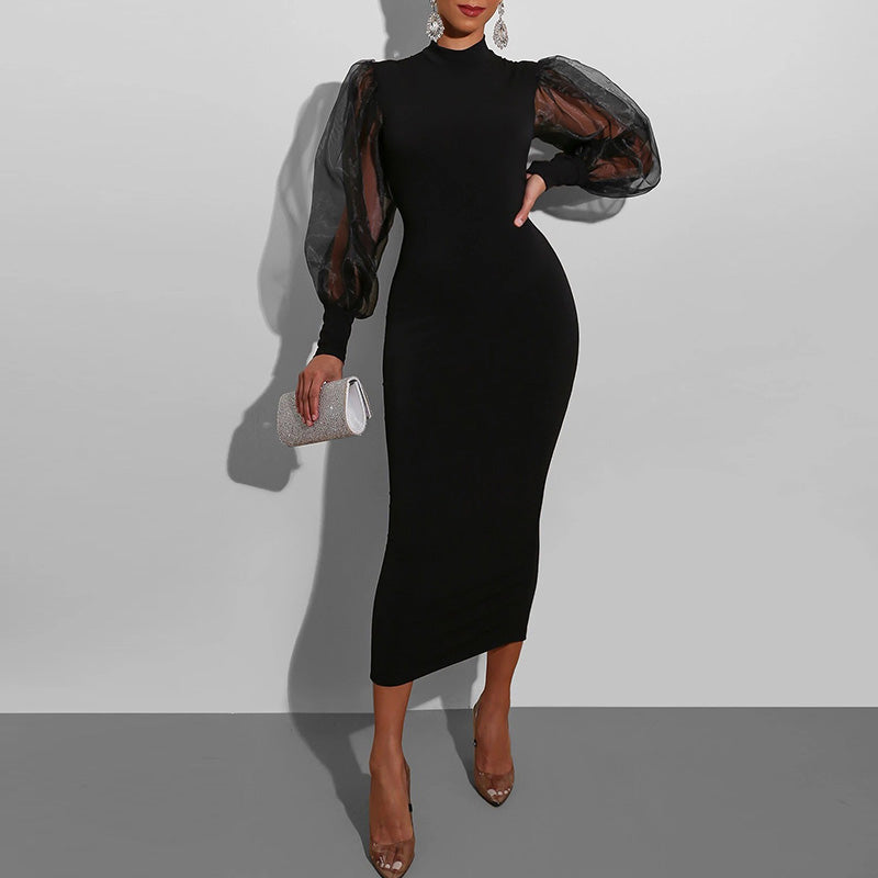 A woman in a black midi dress with sheer puff sleeves and a mock neck, holding a silver clutch.