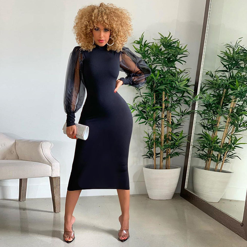 A woman with curly blonde hair wearing a black midi dress with sheer puff sleeves and a mock neck, posing in a room with plants and a mirror.