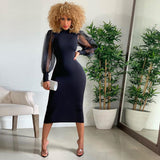 A woman with curly blonde hair wearing a black midi dress with sheer puff sleeves and a mock neck, posing in a room with plants and a mirror.