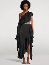 A woman wears a black one-shoulder ruffled dress with a cut-out and high-low hem, standing against a white background.