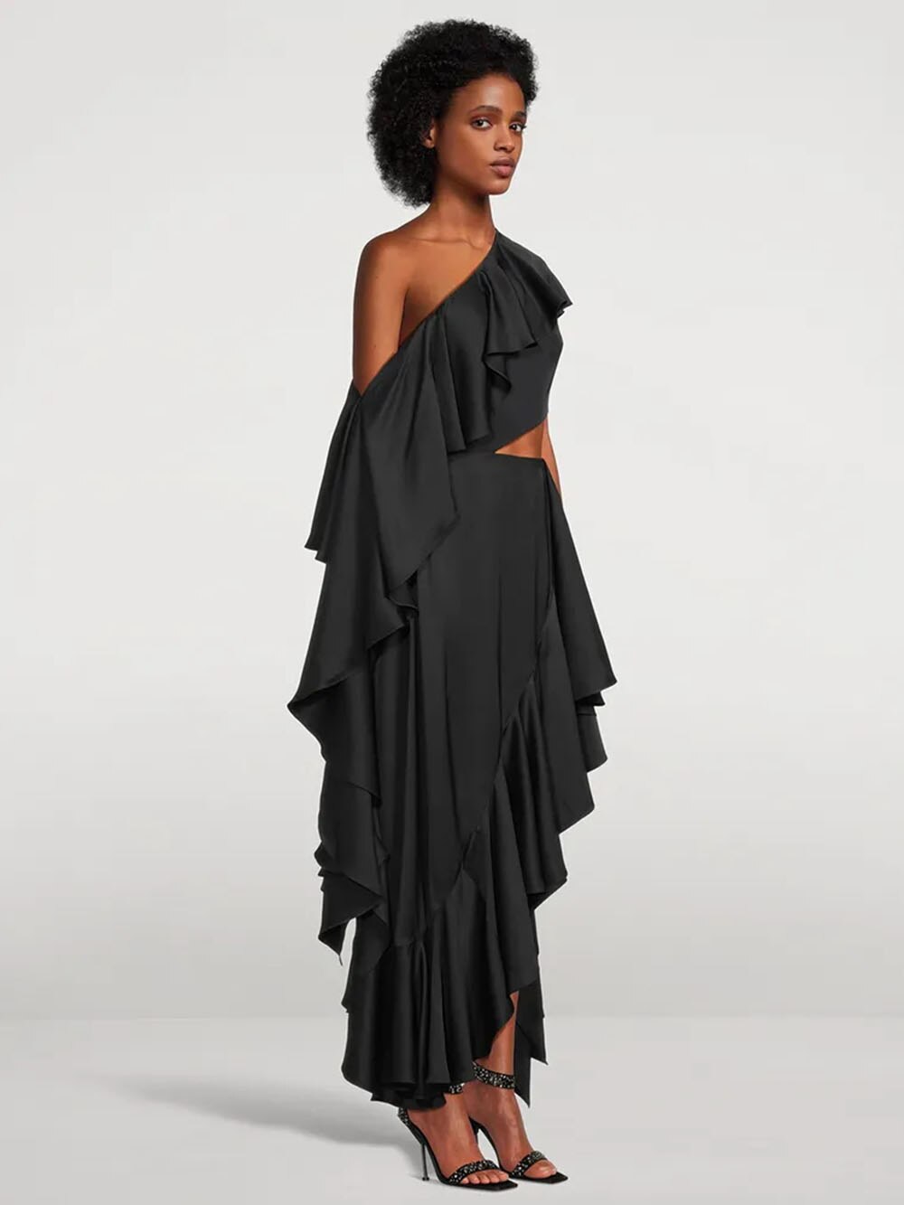 A woman in a black one-shoulder ruffled dress with a cut-out and high-low hem, shown from the side.