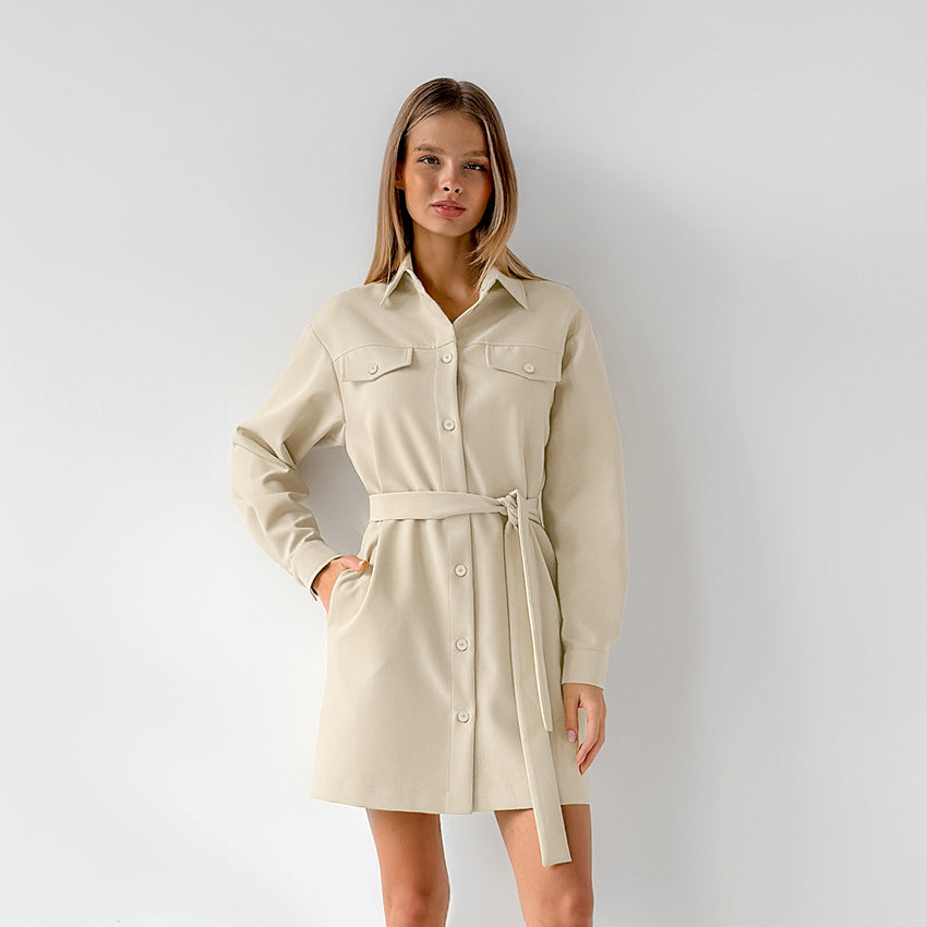 Woman wearing a long-sleeved beige shirt dress with a belted waist and chest pockets, with one hand in her pocket, standing against a white wall.