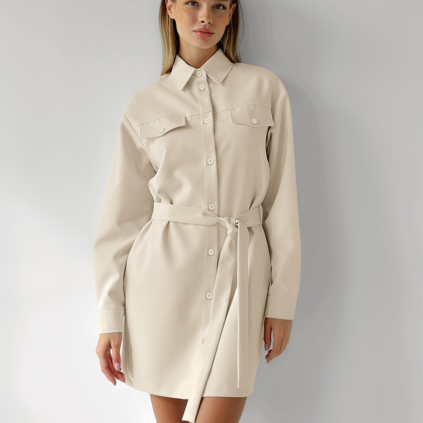 Woman wearing a long-sleeved beige shirt dress with a belted waist and chest pockets, standing against a white wall.