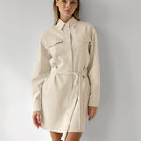 Woman wearing a long-sleeved beige shirt dress with a belted waist and chest pockets, standing against a white wall.