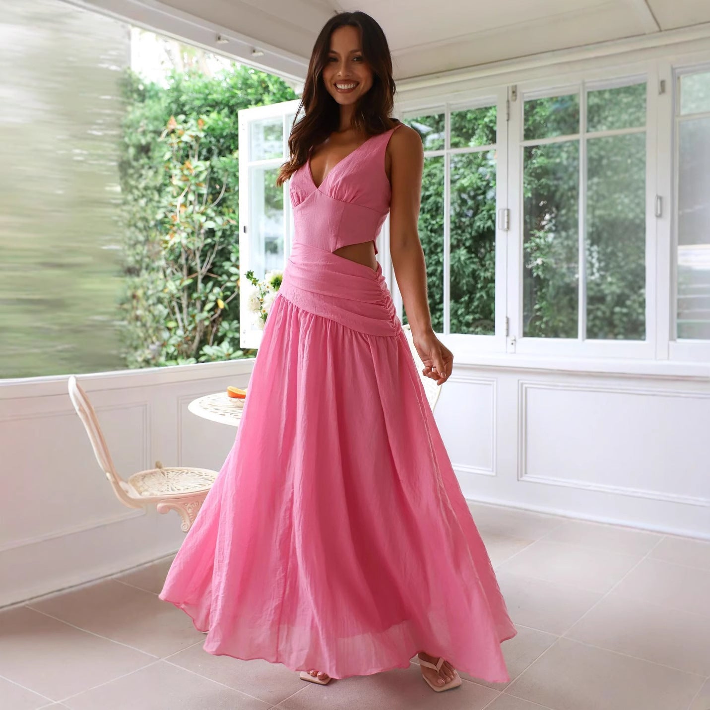 A woman in a pink maxi dress with a cut-out waist and V-neckline stands in a sunlit room with large windows.