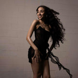 A woman with long wavy dark hair smiling and looking to the side, wearing a black strapless corset dress with sheer fabric and a draped skirt.