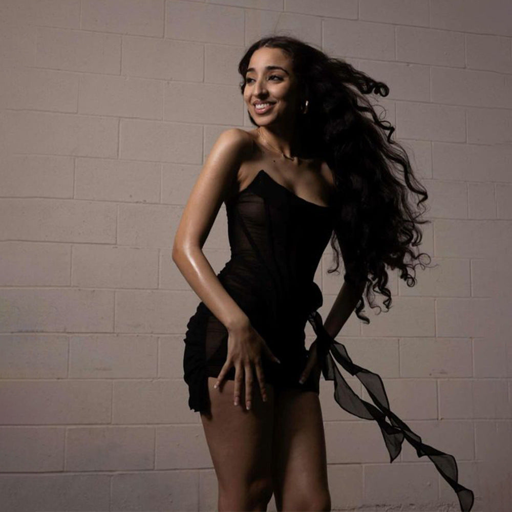 A woman with long wavy dark hair smiling and looking to the side, wearing a black strapless corset dress with sheer fabric and a draped skirt.