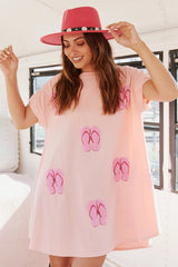 A woman wearing a pink t-shirt dress with sequined flip-flop prints and a pink hat, smiling.