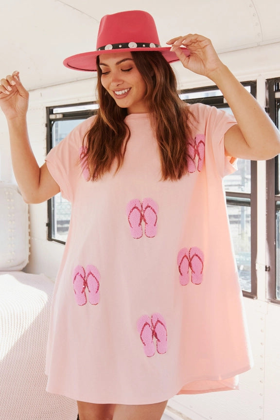 A woman wearing a pink t-shirt dress with sequined flip-flop prints and a pink hat, smiling.