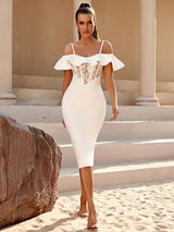 A woman in a white off-the-shoulder midi dress with a lace bust panel walks towards the camera on a sandy surface, with stone columns in the background.