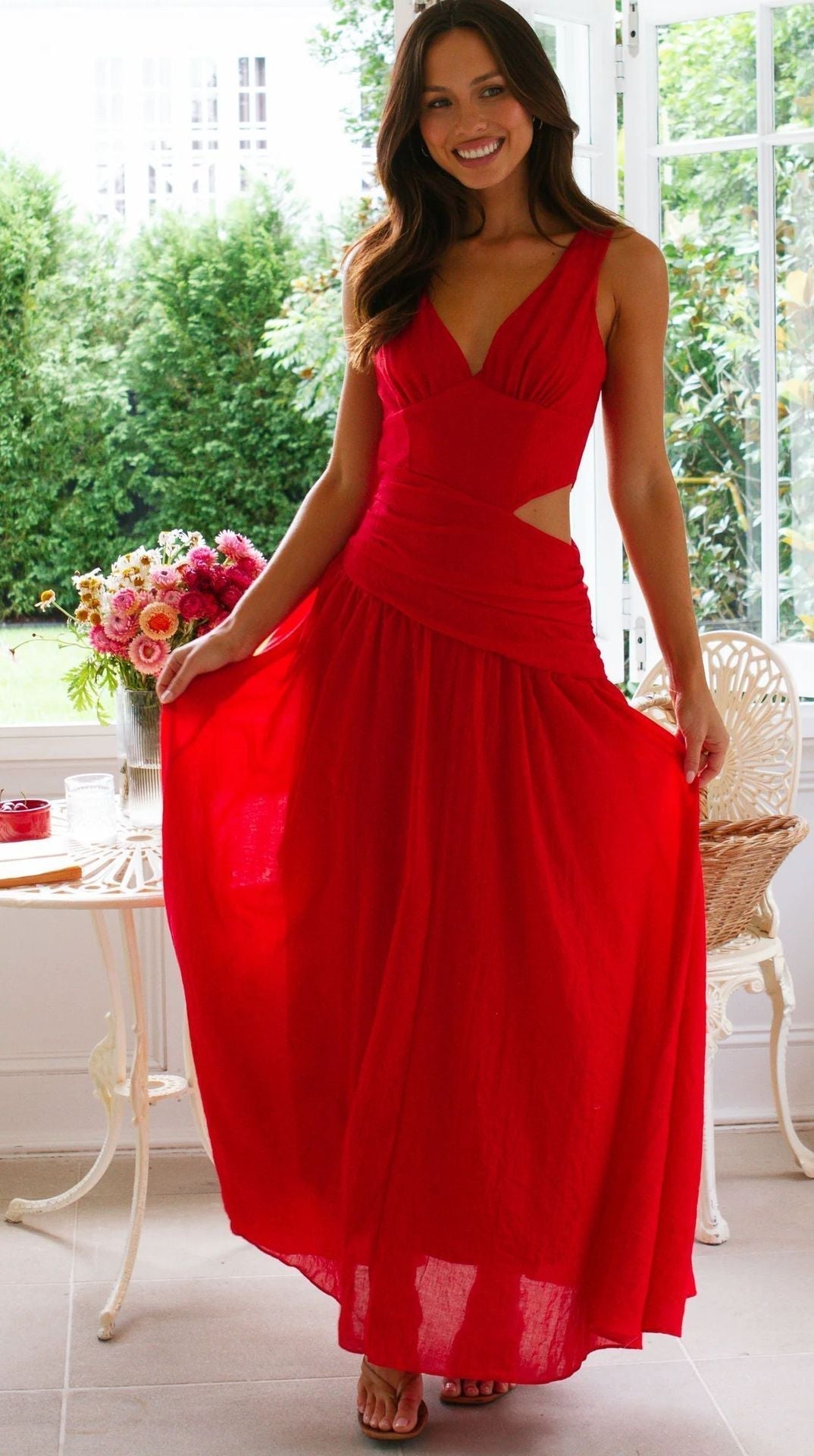 A woman in a red maxi dress with a cut-out waist and V-neckline stands by a table with a vase of flowers.