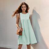 A young woman stands against a light-colored wall, wearing a mint green sleeveless A-line dress with a button-front and pleated yoke. She has a brown crossbody bag and her hair is blowing in the wind.