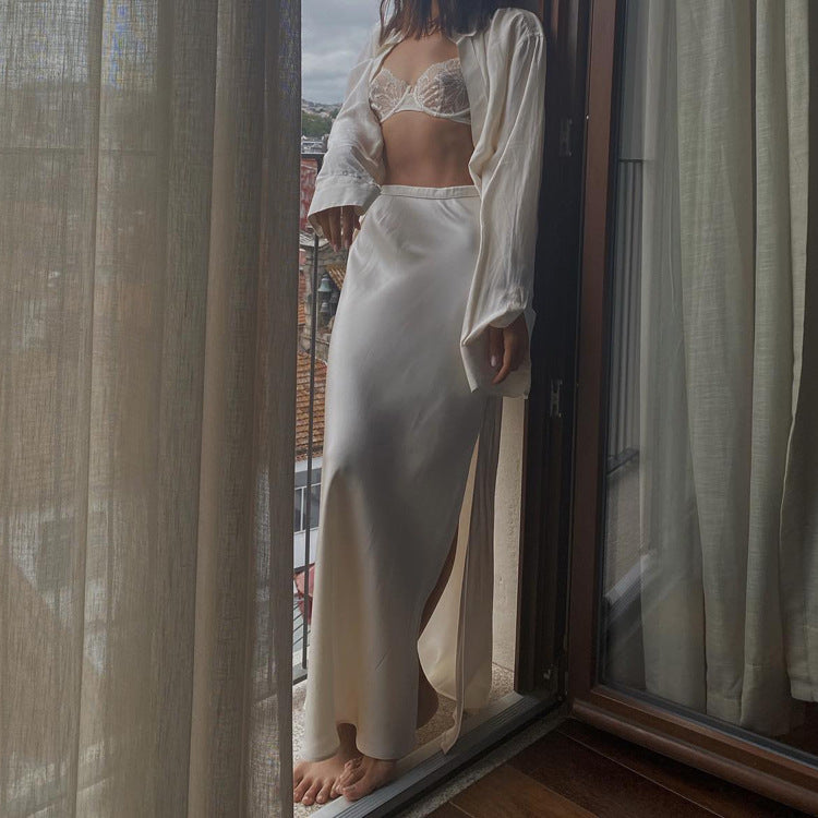 A woman stands on a balcony wearing a white satin maxi skirt, matching shirt, and a lace bralette.