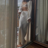 A woman stands on a balcony wearing a white satin maxi skirt, matching shirt, and a lace bralette.