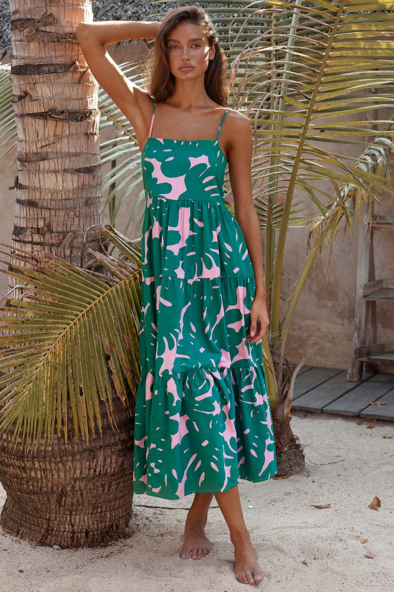 woman wearing a green and pink leaf print tiered midi dress leaning against a palm tree