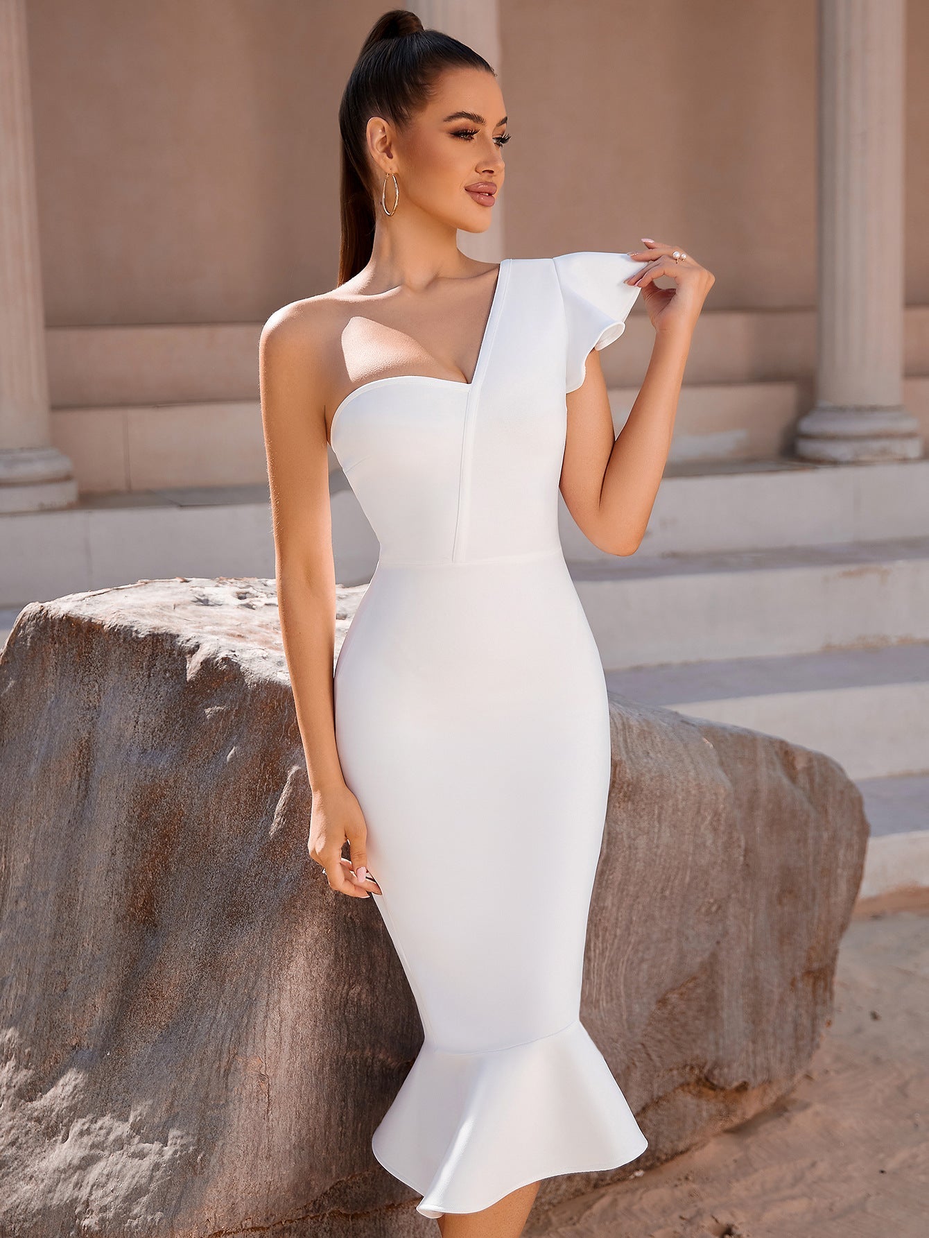 A woman in a white one-shoulder dress with a ruffled sleeve and a mermaid hemline poses next to a large rock.