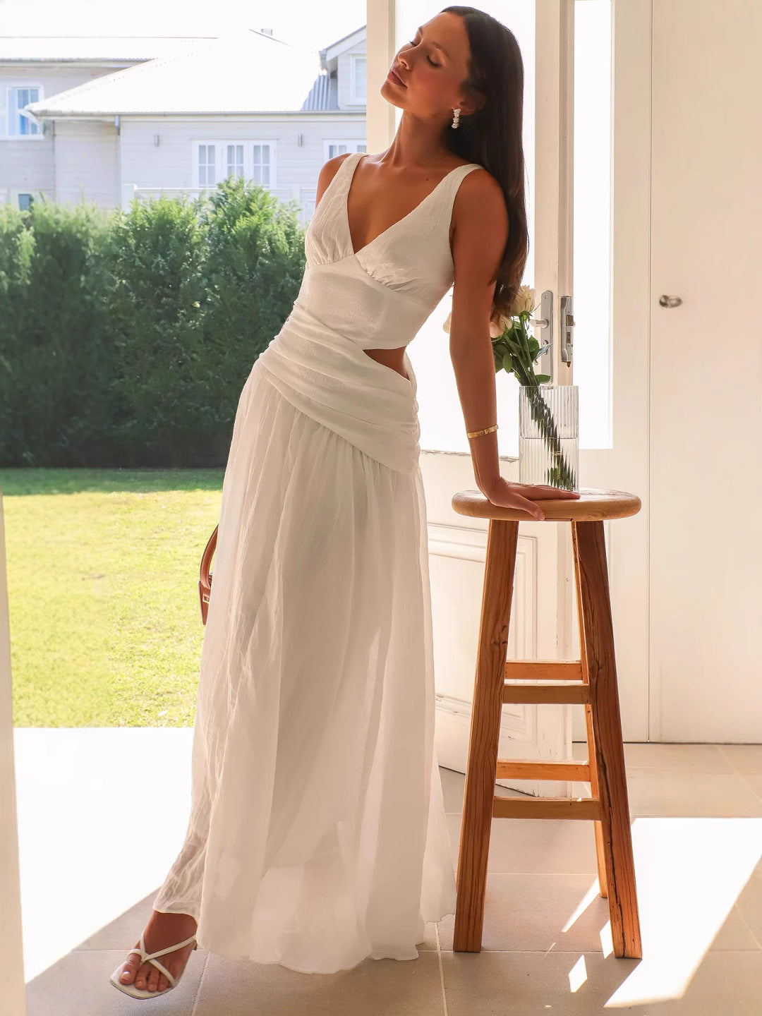 A woman in a white maxi dress with a cut-out waist and V-neckline leans against a wooden stool.
