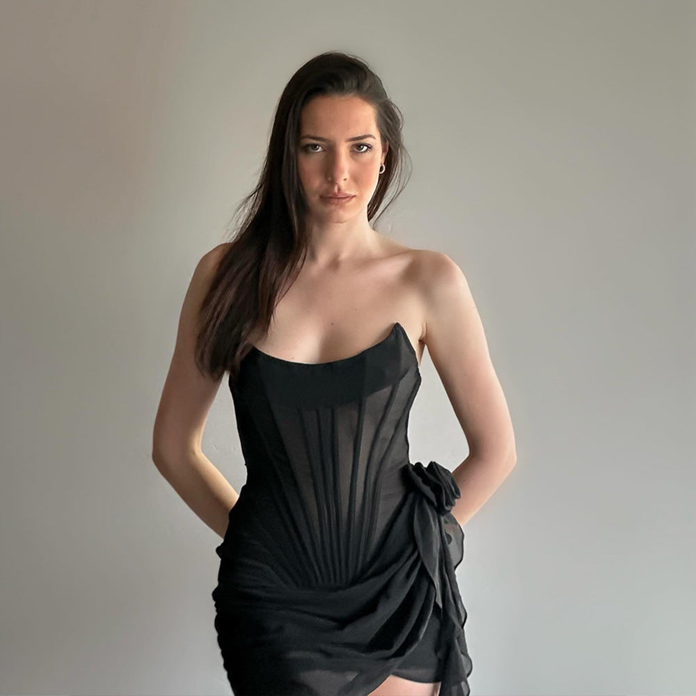 A woman in a black strapless corset dress with sheer fabric and a draped skirt, with her hands behind her back, looking at the camera.