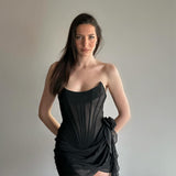 A woman in a black strapless corset dress with sheer fabric and a draped skirt, with her hands behind her back, looking at the camera.